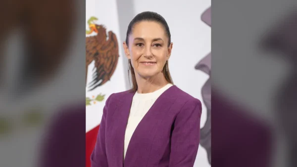 Una mujer en un podio, hablando ante un micrófono, con un fondo de banderas de México.